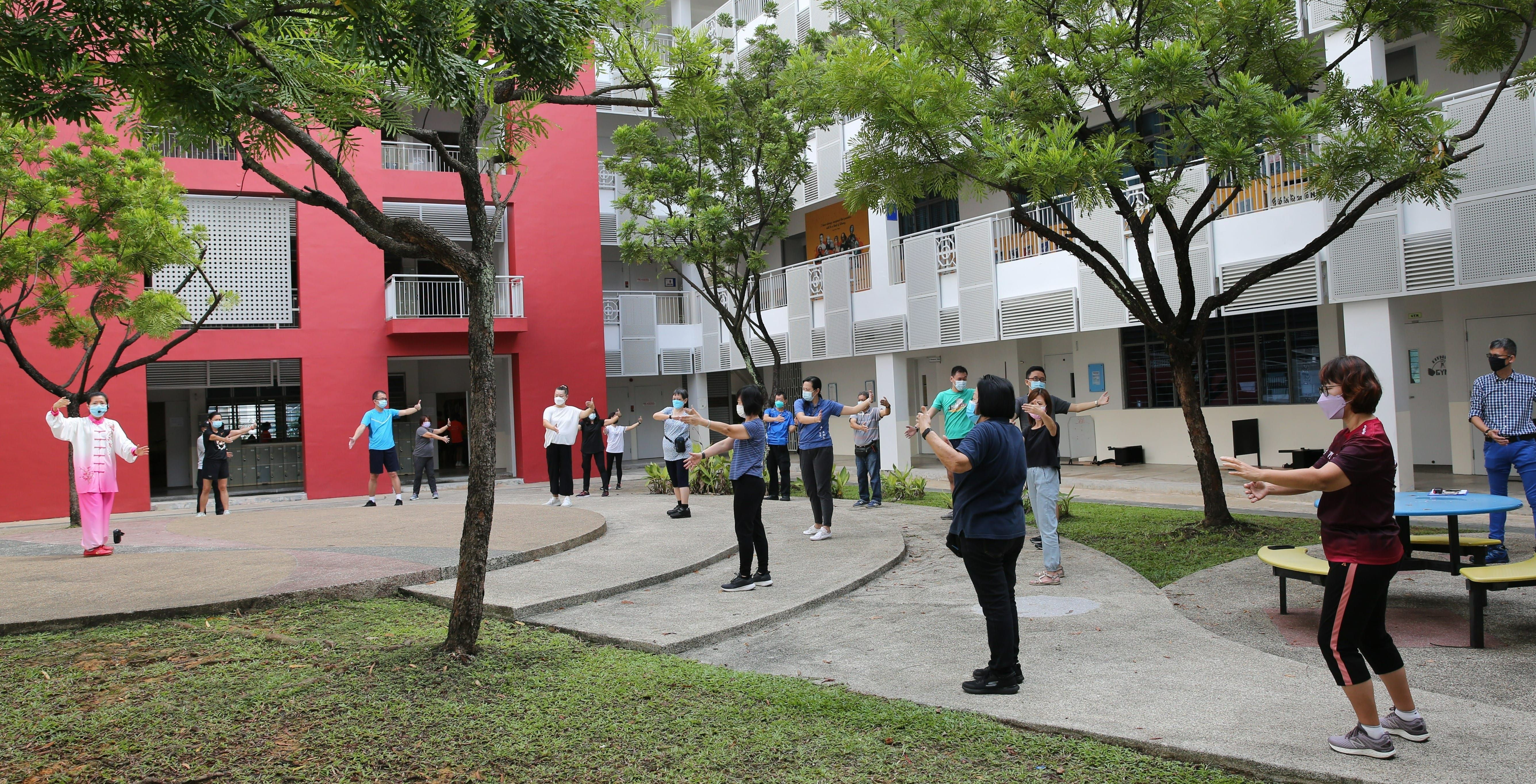 Masked group doing outdoor exercise with arms raised in a courtyard beside red and white buildings.